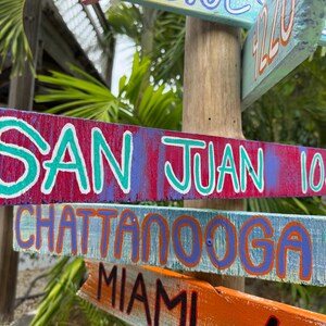 May include: Wooden directional signs with painted text. The top sign reads "SAN JUAN 1030" in white and teal lettering on a red and purple background. Other signs point to Chattanooga, Miami, and more.
