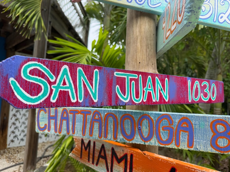 May include: Wooden directional signs with painted text. The top sign reads "SAN JUAN 1030" in white and teal lettering on a red and purple background. Other signs point to Chattanooga, Miami, and more.