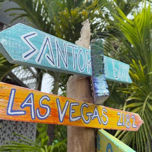 May include: Wooden directional signs point towards Santorini, Las Vegas, and other destinations. The signs are painted in bright colors, including blue, orange, and yellow, with white lettering. The signpost is made of wood and is surrounded by lush green foliage.
