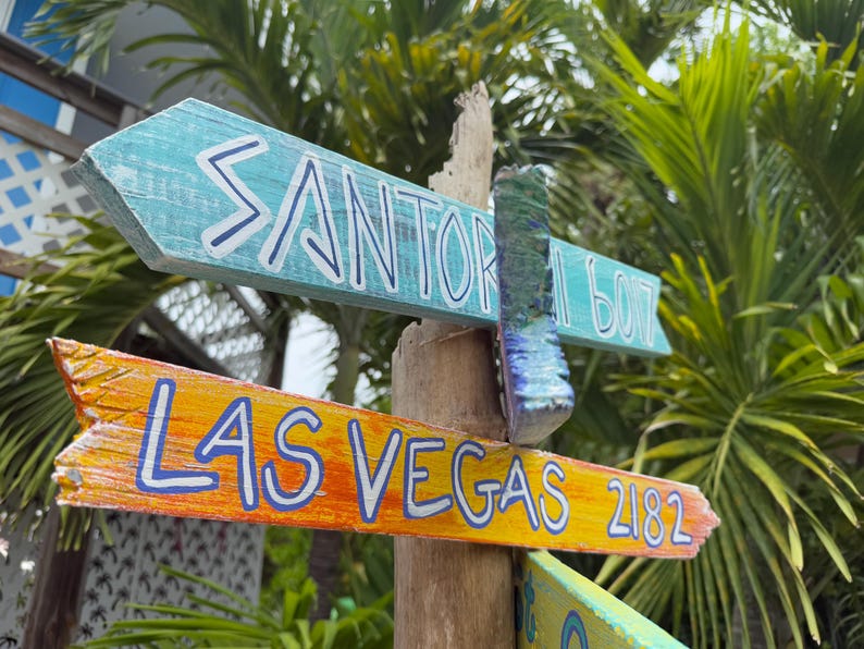 May include: Wooden directional signs point towards Santorini, Las Vegas, and other destinations. The signs are painted in bright colors, including blue, orange, and yellow, with white lettering. The signpost is made of wood and is surrounded by lush green foliage.