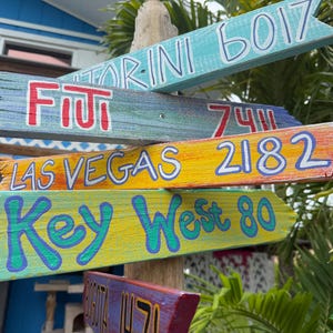 May include: A collection of colorful wooden directional signs. The signs point in various directions, with destinations like Fiji, Las Vegas, and Key West. Each sign is painted with a different color and features the destination name and a distance.