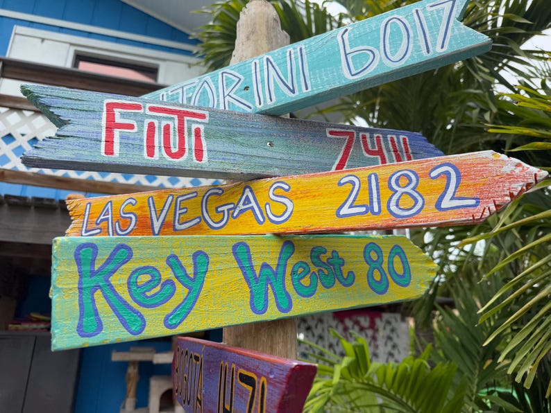 May include: A collection of colorful wooden directional signs. The signs point in various directions, with destinations like Fiji, Las Vegas, and Key West. Each sign is painted with a different color and features the destination name and a distance.