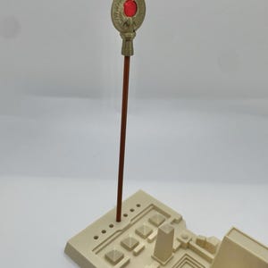 May include: A beige model of an ancient structure with a brown staff topped with a gold-colored emblem and a red gem. The base features pyramid and rectangular shapes.
