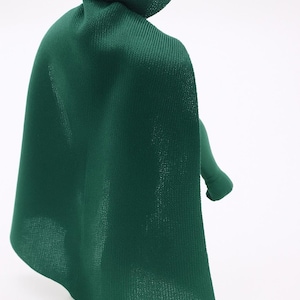 Pode incluir: Uma capa de tecido verde com gola alta, drapeada sobre uma figura com cabeça vermelha e listra amarela. O braço da figura é visível, também em verde. A capa tem uma aparência texturizada.