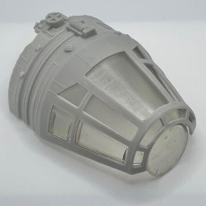 May include: A gray plastic model of a spaceship cockpit. The model has a cylindrical shape with detailed exterior features, including panels, windows, and a circular front section. The cockpit windows are clear.
