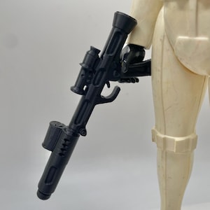 May include: A vintage Stormtrooper action figure, featuring a black blaster rifle. The figure is primarily off-white with black accents, showcasing the iconic design from the Star Wars franchise. The blaster is held in the figure's right hand.