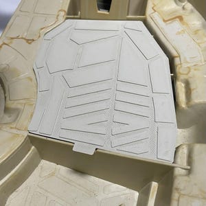 以下が含まれることがあります： ベージュ色のプラスチック製玩具車両部品。中央の平らなパネルには、長方形や台形の形状が浮き彫りになった幾何学模様が施されています。周囲の構造には成形されたグリッド線と円筒形のペグがあります。