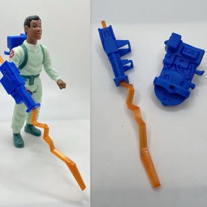 May include: A Ghostbusters action figure in a light green jumpsuit with a blue proton pack and a blue and orange proton gun. The figure has a brown complexion and is posed with the proton gun.