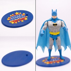 May include: A Batman action figure with a light blue cape, grey suit, yellow utility belt, and black bat emblem. The figure stands on a blue oval display base featuring the text "SUPER POWERS" surrounded by yellow stars and red bursts.