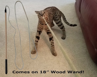 May include: A brown and tan tabby cat plays with a long, white string toy attached to a wooden wand. The wand is 18 inches long and has a black handle. The text "Comes on 18" Wood Wand!" is printed on the floor below the cat.