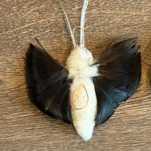 May include: A decorative ornament shaped like a moth or butterfly, featuring a central body made of white fuzzy material, black feathers extending as wings, two thin antennae made of white feathers, and a small orange thread loop for hanging.