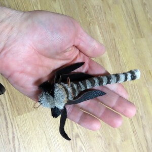 Może przedstawiać: Ręcznie robiona zabawka dla kota w czarno-białe paski z futrzastą głową i sk&oacute;rzanymi skrzydłami. Zabawka jest trzymana w dłoni osoby.