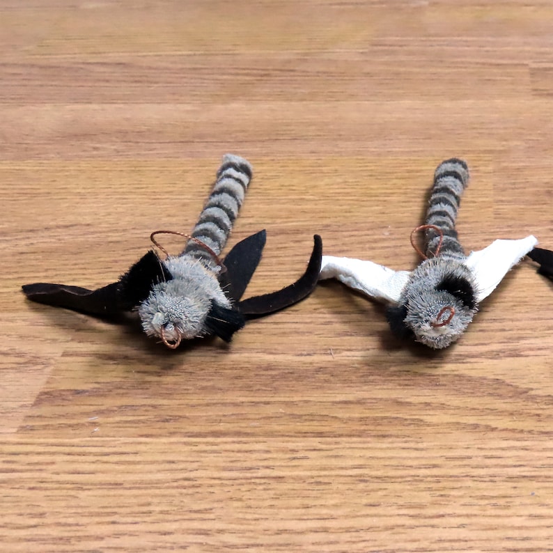 Może przedstawiać: Dwie pluszowe zabawki w kształcie nietoperzy z czarnymi skrzydłami i szaro-białymi paskowanymi ciałami. Zabawki mają miedziane akcenty z drutu wok&oacute;ł głowy.