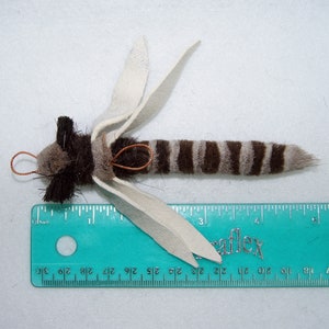 Może przedstawiać: Ręcznie robiona zabawka dla kota z brązowo-białym paskowanym ogonem, brązowym futrem i białymi sk&oacute;rzanymi skrzydłami. Zabawka ma około 10 cm długości i jest mierzona linijką.