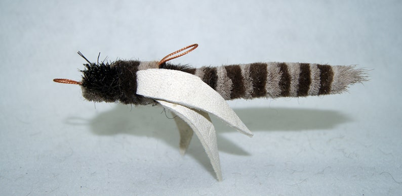 Może przedstawiać: Pluszowa zabawka w brązowo-białe paski z białymi skrzydłami i miedzianymi antenami z drutu. Zabawka jest zaprojektowana tak, aby przypominała muchę.