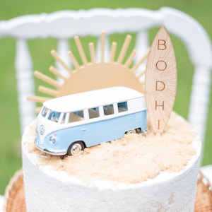 May include: A white cake with a blue and white toy van, a gold sun, and a wooden surfboard with the name "BODHI" on it. The cake is decorated with a sandy brown frosting.