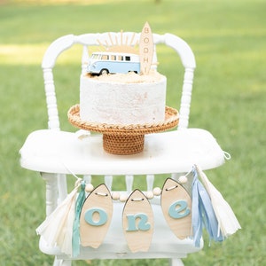 May include: A white high chair with a blue and white surfboard banner that says "one". A cake with a blue and white van and a surfboard topper that says "Bodhi" is on the chair.