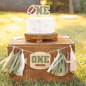 May include: A white cake with a football and the word "ONE" on top. The cake is on a wooden cake stand and a wooden box with a football-shaped banner that says "ONE" and green and brown tassels.