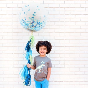 May include: A young boy wearing a gray t-shirt with a dinosaur graphic and blue jeans is standing in front of a white brick wall. He is holding a large blue and green confetti balloon with a blue and green tassel.