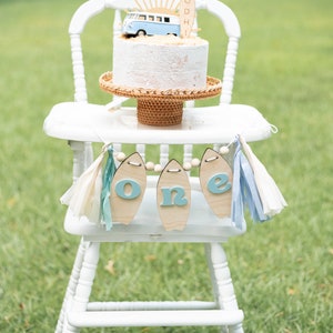 May include: A white high chair with a white cake decorated with a blue and white toy van and a wooden surfboard with the name "BODHI" on it. A blue and white banner with the word "one" on it hangs from the chair.