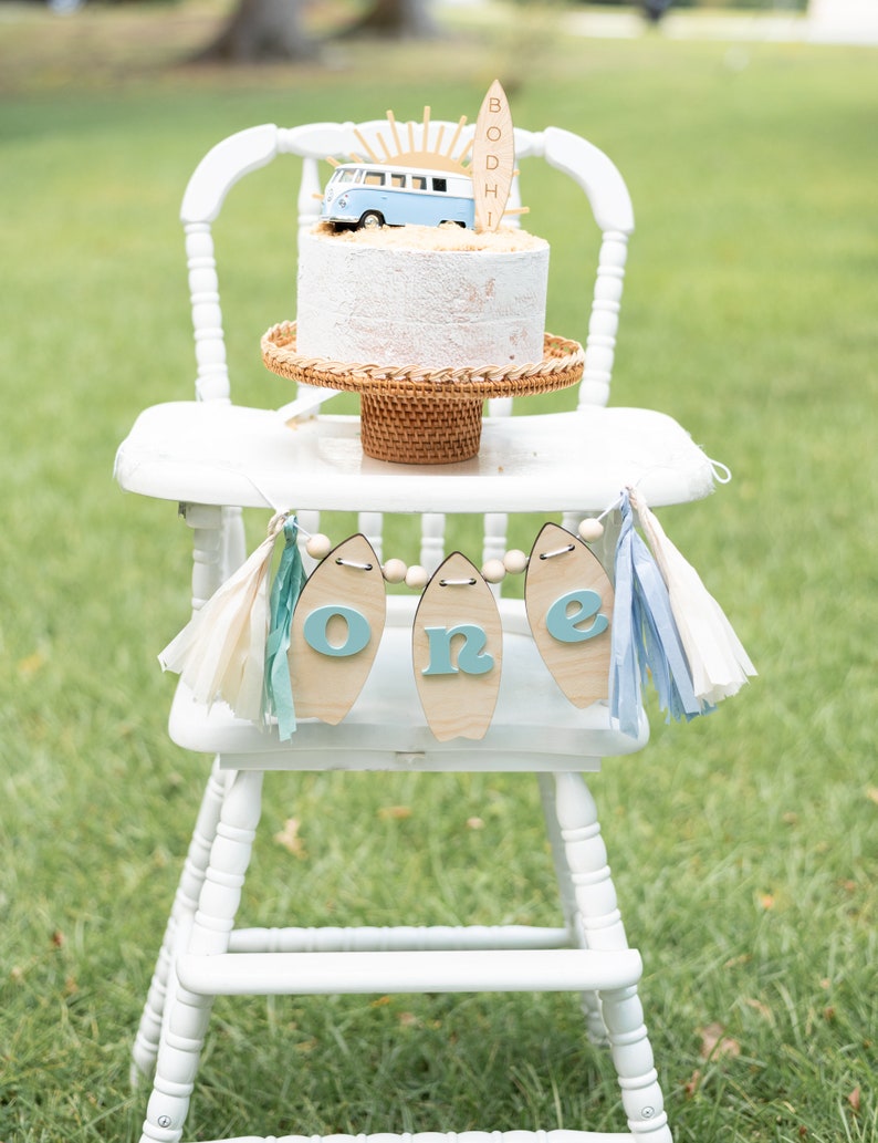 May include: A white high chair with a white cake decorated with a blue and white toy van and a wooden surfboard with the name "BODHI" on it. A blue and white banner with the word "one" on it hangs from the chair.