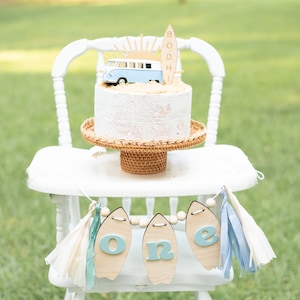 May include: A white high chair with a blue and white cake decorated with a surfboard, a sun, and a blue van. The cake is on a wicker cake stand. A blue and white banner with the word "one" on it hangs from the chair.