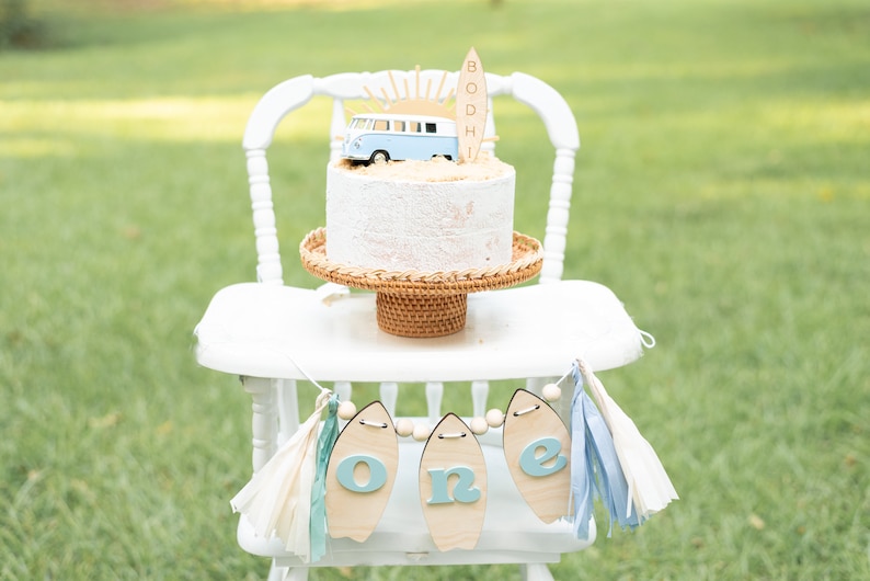 May include: A white high chair with a blue and white cake decorated with a surfboard, a sun, and a blue van. The cake is on a wicker cake stand. A blue and white banner with the word "one" on it hangs from the chair.