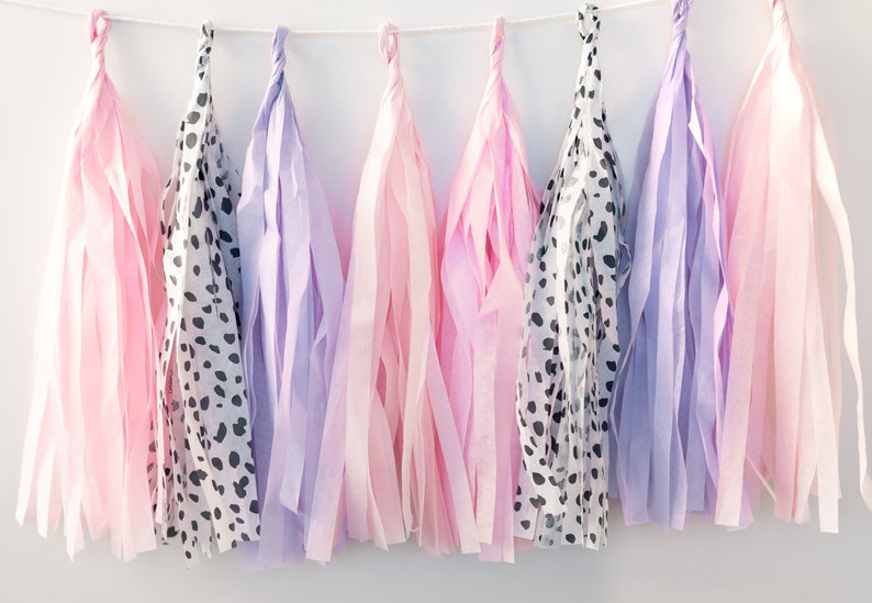 Puede incluir: Una guirnalda de borlas de papel de seda en rosa, morado y con lunares negros y blancos. Las borlas est&aacute;n enhebradas con una cuerda blanca.