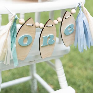 May include: A white high chair with a blue and white surfboard-shaped banner that says "one" hanging from it. The banner is decorated with wooden beads and blue and white tassels.