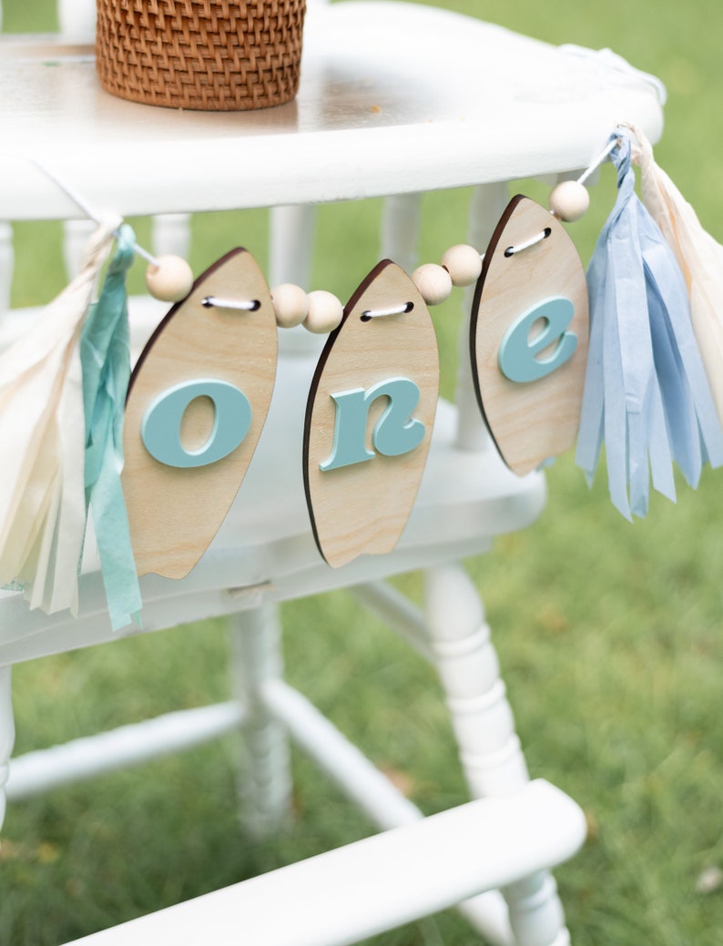 May include: A white high chair with a blue and white surfboard-shaped banner that says "one" hanging from it. The banner is decorated with wooden beads and blue and white tassels.