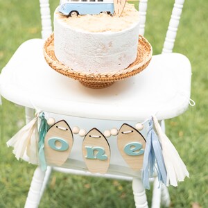 May include: A white high chair with a wooden banner that says "one" in blue letters. The banner is strung with wooden beads and has blue and white tassels. A white cake with a blue and white camper van and a surfboard that says "Bodhi" is on the chair.