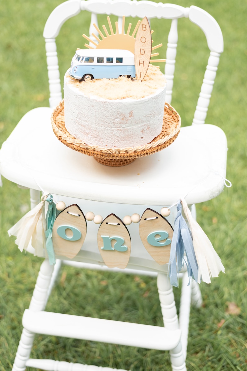May include: A white high chair with a wooden banner that says "one" in blue letters. The banner is strung with wooden beads and has blue and white tassels. A white cake with a blue and white camper van and a surfboard that says "Bodhi" is on the chair.