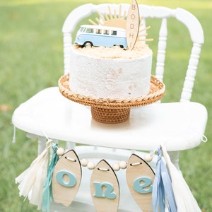 May include: A white high chair with a white cake decorated with a blue and white toy van, a wooden surfboard with the name "BODHI" on it, and a yellow sun. A banner with the word "one" on it hangs from the chair. The banner is made of wooden surfboards with blue letters and white beads.