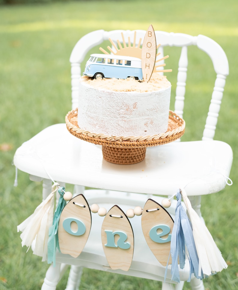 May include: A white high chair with a white cake decorated with a blue and white toy van, a wooden surfboard with the name "BODHI" on it, and a yellow sun. A banner with the word "one" on it hangs from the chair. The banner is made of wooden surfboards with blue letters and white beads.