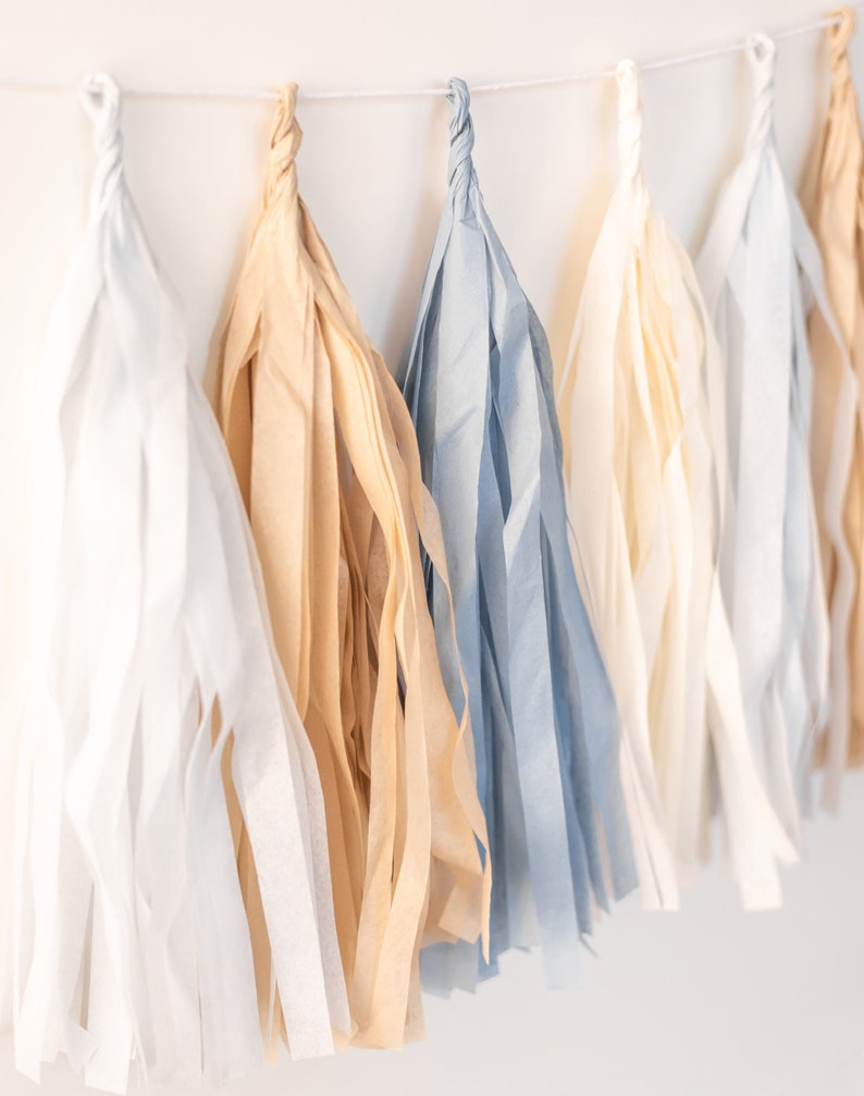 Puede incluir: Una guirnalda de borlas de papel de seda en colores blanco, beige y azul claro. Las borlas cuelgan de una cuerda blanca.