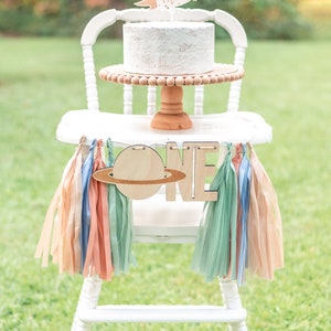 May include: White high chair with a white cake decorated with a rocket and the name Liam. The cake is on a wooden cake stand. A wooden banner with the word "ONE" and a planet is hanging from the chair. The banner is decorated with pastel colored tissue paper tassels.