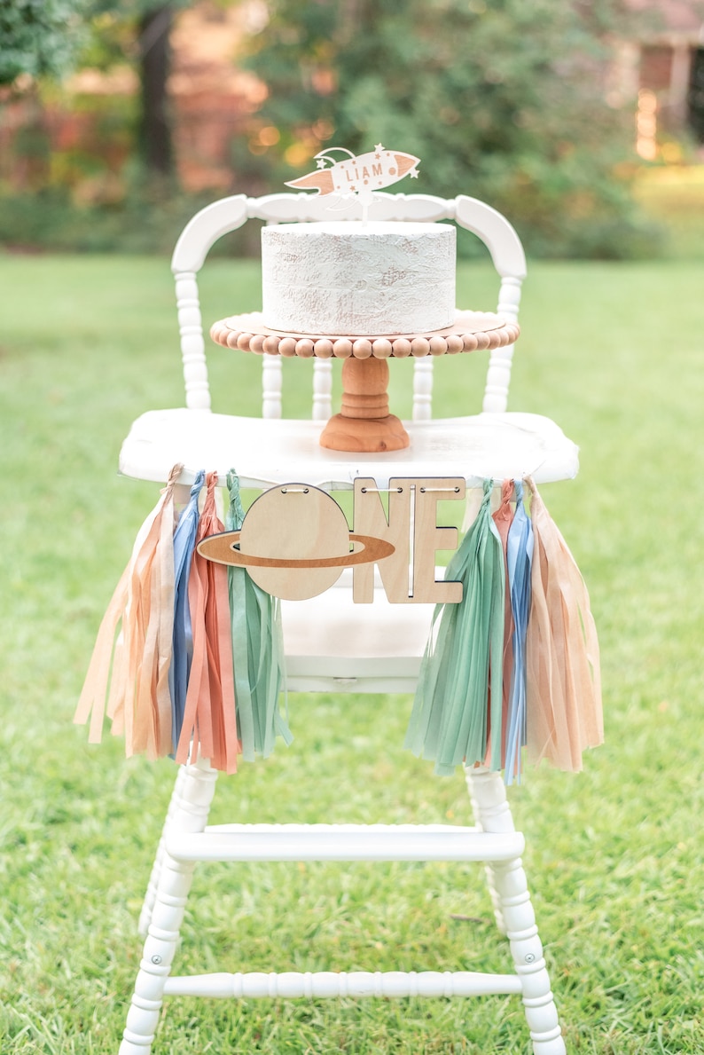 May include: White high chair with a white cake decorated with a rocket and the name Liam. The cake is on a wooden cake stand. A wooden banner with the word "ONE" and a planet is hanging from the chair. The banner is decorated with pastel colored tissue paper tassels.