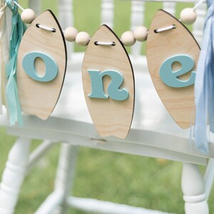 May include: A wooden banner with the word "one" spelled out on it. Each letter is on a separate surfboard-shaped piece of wood. The banner is strung together with wooden beads and blue and white ribbon.