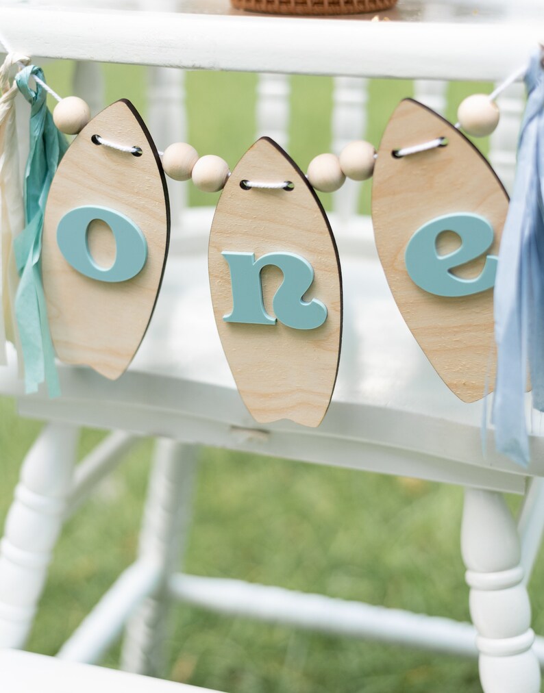 May include: A wooden banner with the word "one" spelled out on it. Each letter is on a separate surfboard-shaped piece of wood. The banner is strung together with wooden beads and blue and white ribbon.