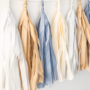 Puede incluir: Una guirnalda de borlas de papel de seda en blanco, beige y azul. Las borlas est&aacute;n unidas con una cuerda blanca.
