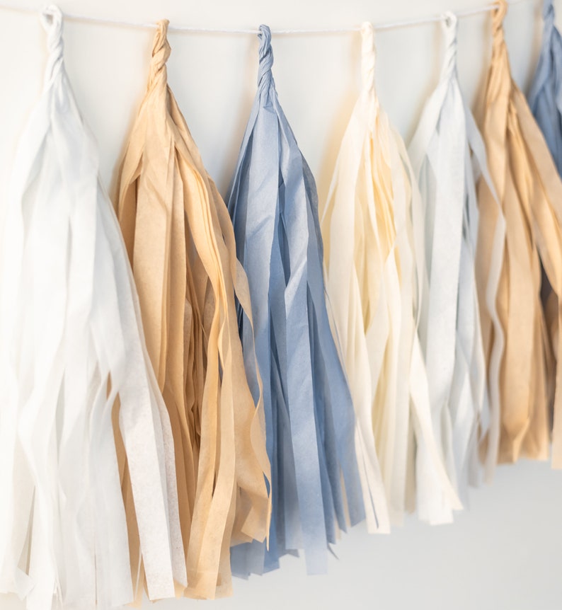 Puede incluir: Una guirnalda de borlas de papel de seda en blanco, beige y azul. Las borlas est&aacute;n unidas con una cuerda blanca.