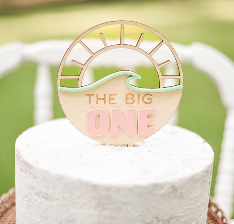 May include: A wooden cake topper with a circular design featuring a sun and wave, with the text "THE BIG ONE" in pink letters. The topper is placed on top of a white frosted cake, perfect for a birthday celebration.