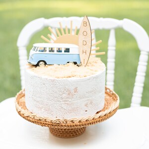 May include: A white cake with a blue and white toy van, a wooden surfboard with the name "BODHI" on it, and a brown sugar-like topping. The cake is on a brown wicker cake stand.