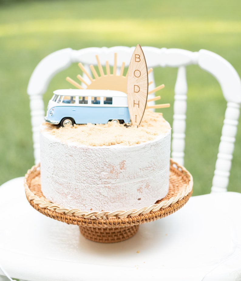 May include: A white cake with a blue and white toy van, a wooden surfboard with the name "BODHI" on it, and a brown sugar-like topping. The cake is on a brown wicker cake stand.