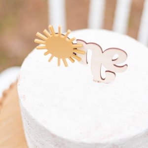 Puede incluir: Un pastel blanco glaseado con un sol amarillo y un adorno de madera con la palabra "uno". El pastel est&aacute; sobre un soporte de pastel de madera.