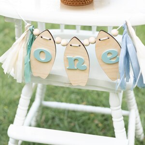 May include: A white high chair with a wooden surfboard banner that says "one" in blue letters. The banner is decorated with wooden beads and blue and white tassels.