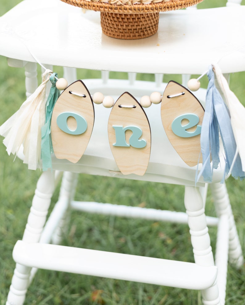 May include: A white high chair with a wooden surfboard banner that says "one" in blue letters. The banner is decorated with wooden beads and blue and white tassels.
