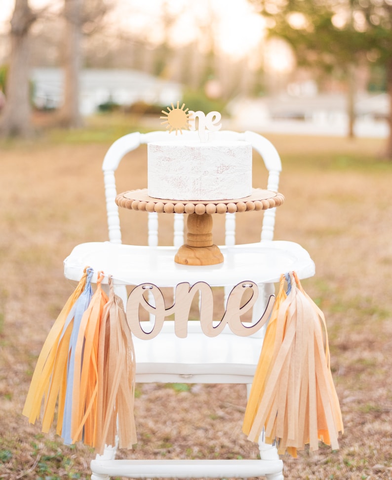 Puede incluir: Silla alta blanca con un pastel blanco sobre un soporte para pastel de madera. El pastel est&aacute; decorado con un sol dorado y la palabra "one". La silla alta est&aacute; decorada con una pancarta que dice "one" y borlas de papel de seda beige y naranja.