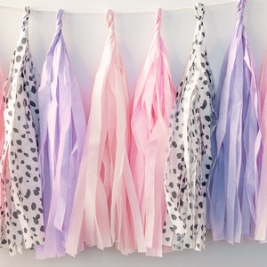 Puede incluir: Una guirnalda de borlas de papel de seda en rosa, morado y un patr&oacute;n de lunares blanco y negro. Las borlas est&aacute;n enhebradas con una cuerda blanca.