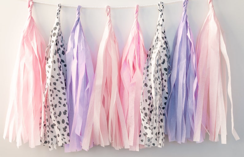 Puede incluir: Una guirnalda de borlas de papel de seda en rosa, morado y un patr&oacute;n de lunares blanco y negro. Las borlas est&aacute;n enhebradas con una cuerda blanca.